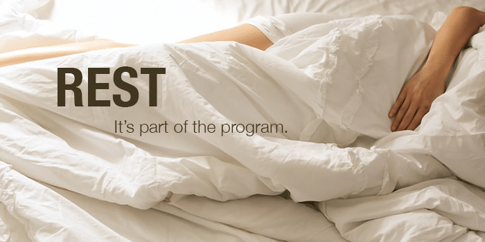 person laying in bed with "REST: it's part of the program" written on the blankets